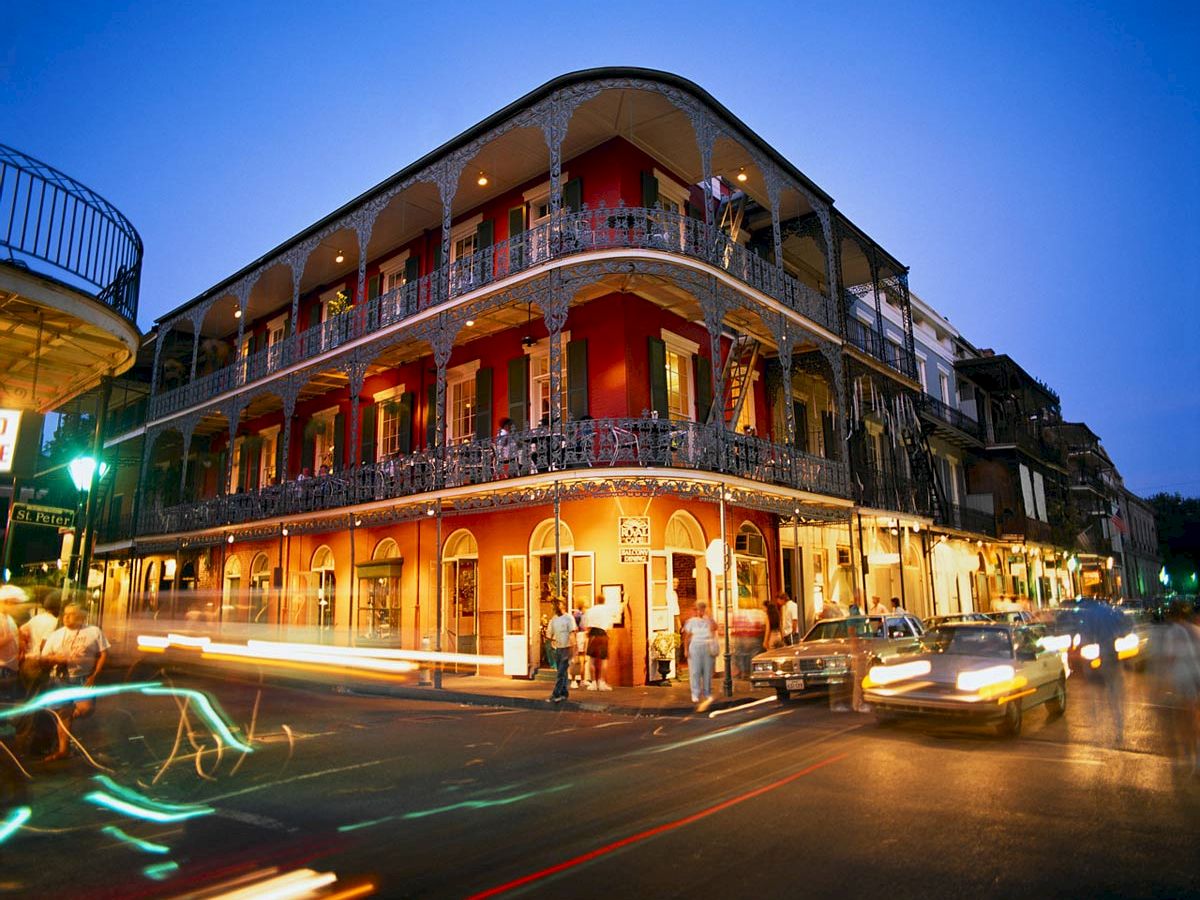 A corner building with intricate ironwork balconies is lit up at dusk, with light trails from passing vehicles on the streets below.