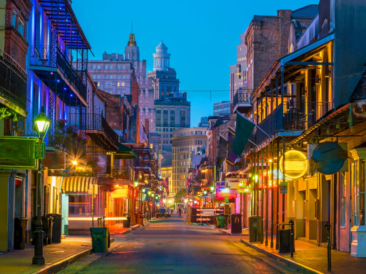 A colorful, lively street with illuminated buildings and shops, likely in a historic district at twilight with city skyscrapers in the background.