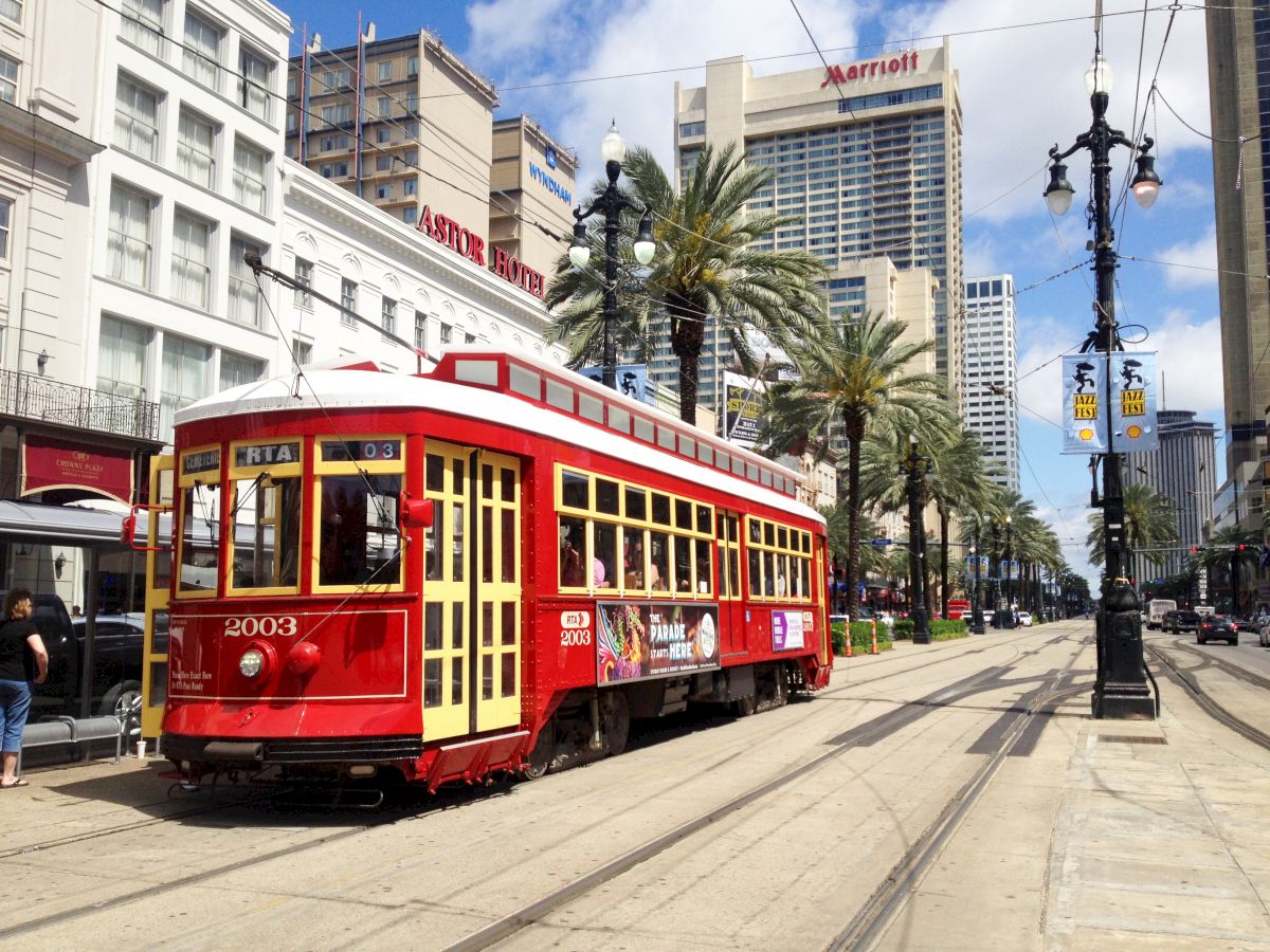 A red streetcar on a track in a city lined with palm trees, tall buildings, and streetlights, under a blue sky with a few clouds.