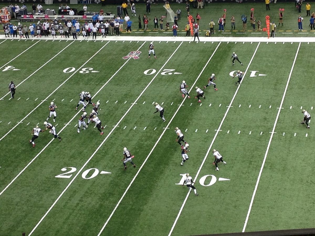 A football game is in progress with players positioned on the field, the ball in play near the 20-yard line, and the crowd visible in the background.