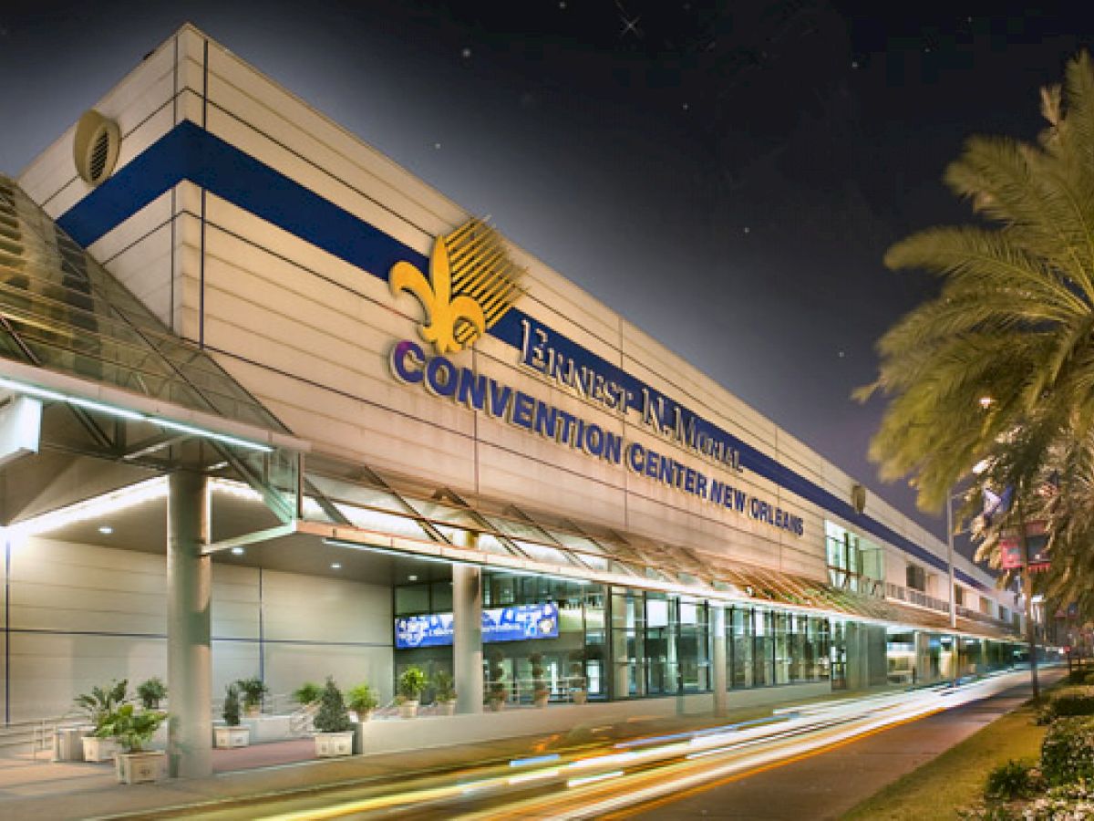 A nighttime view of a large, well-lit building labeled "Ernest N. Morial Convention Center New Orleans," with palm trees on the right side of the image.