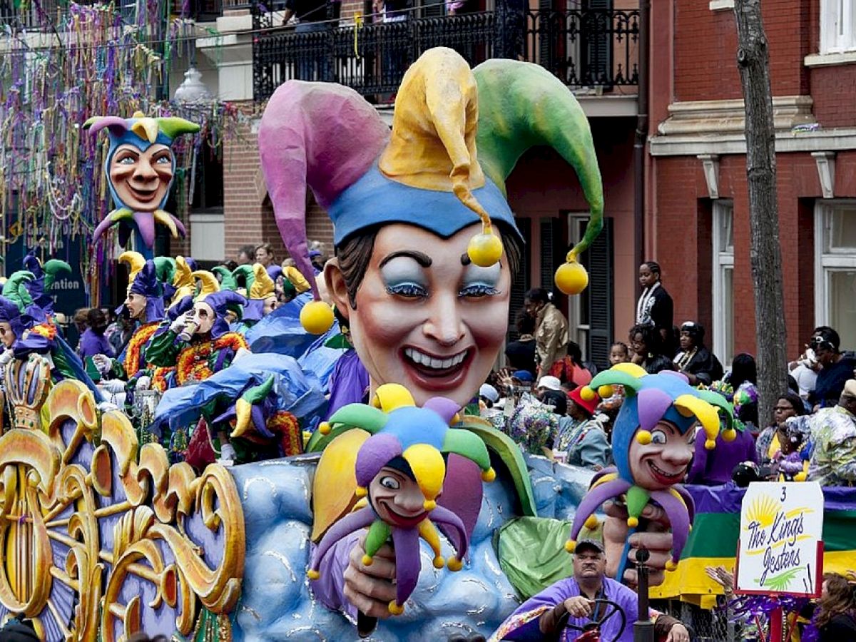 The image shows a vibrant Mardi Gras parade with colorful jester floats, costumed participants, and a cheering crowd on the streets.