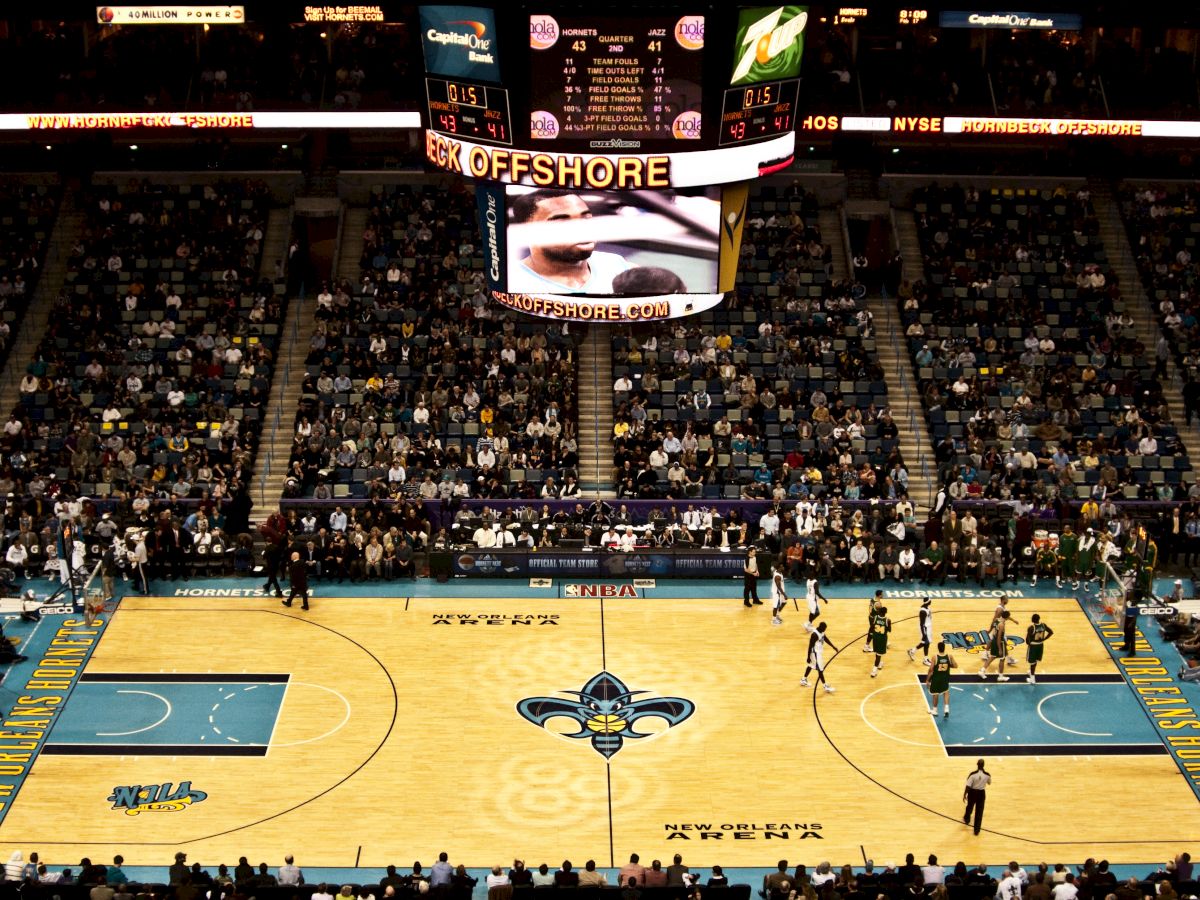 A packed indoor basketball arena with players on the court and a large screen above displaying a game highlight.