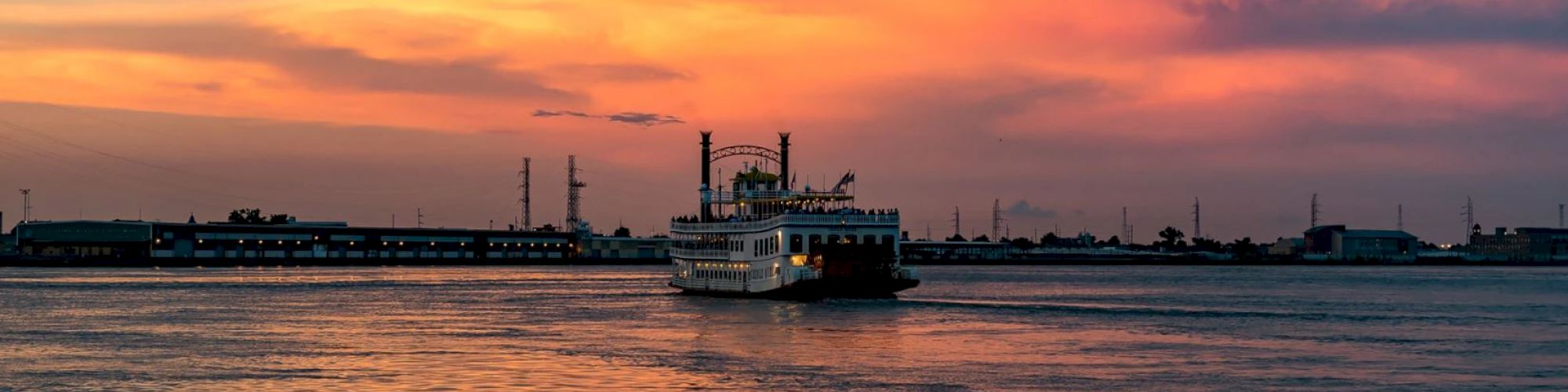 An illuminated riverboat sails across a calm waterway under a vibrant orange and pink sunset sky, with distant buildings lining the horizon.