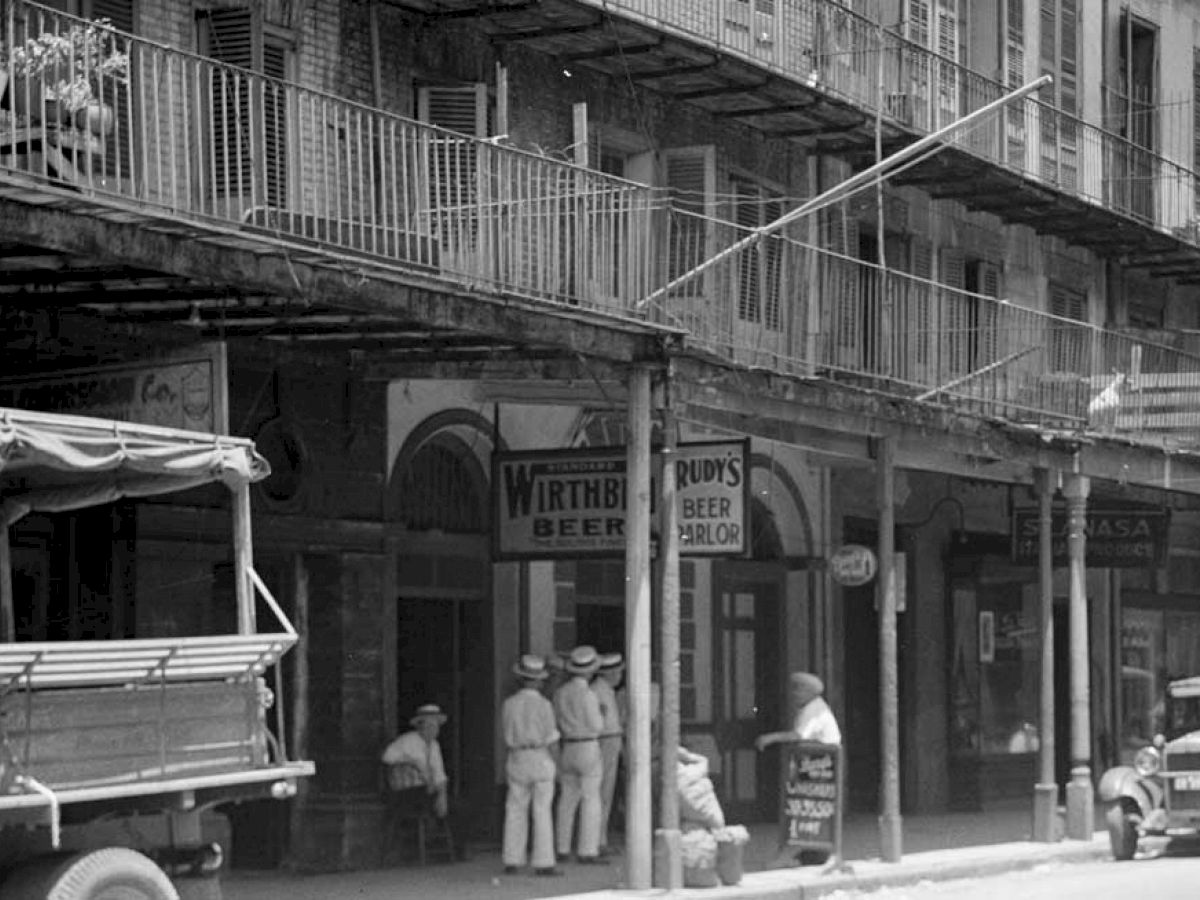 A street scene showing people in white clothing, vintage vehicles, and signs for 