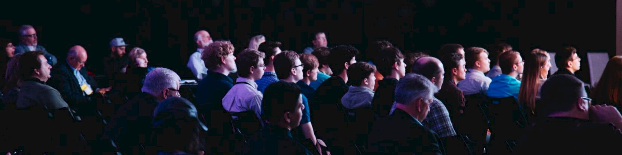 A group of people is seated in rows, attentively listening to a presentation or lecture in a dimly lit room.