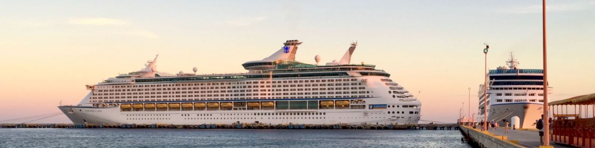 Two large cruise ships docked at a pier at sunset, with calm water and a clear sky in the background.