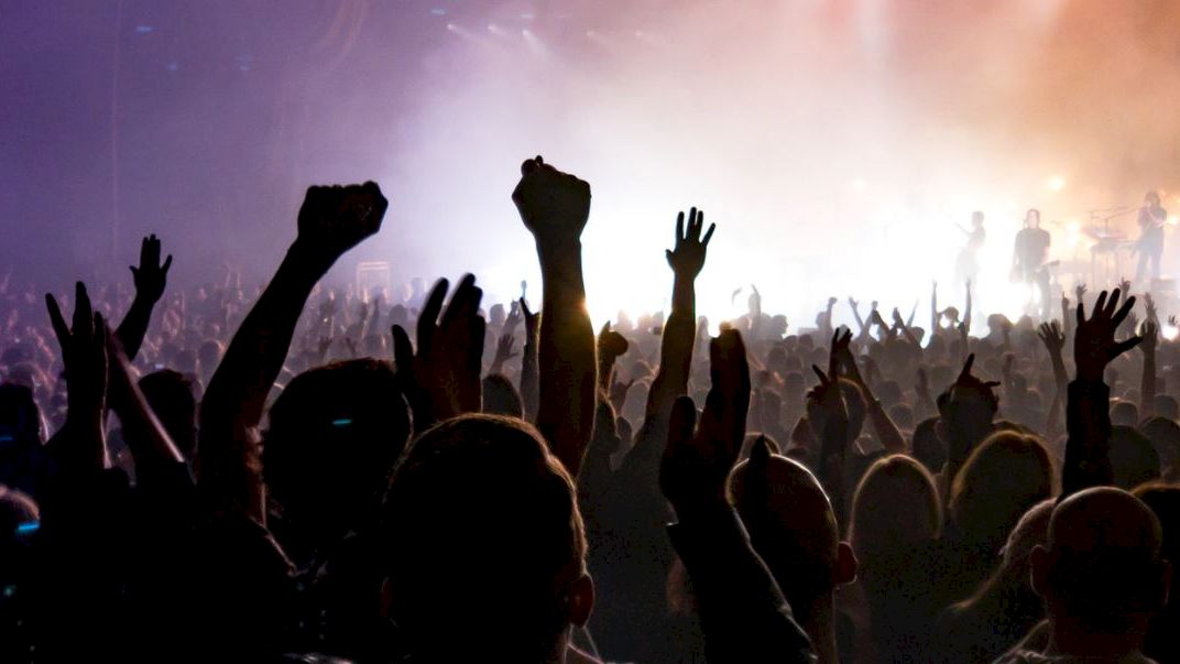 A crowd of people at a concert with their hands raised in the air, lit by colorful stage lights and surrounded by a lively atmosphere.