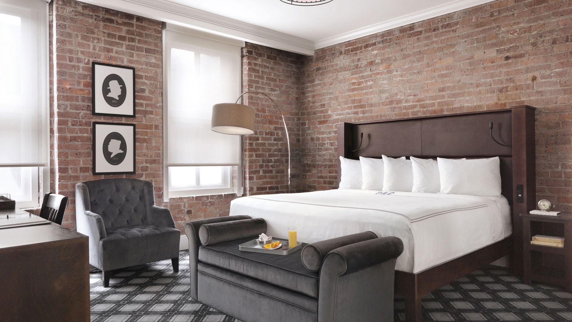 A modern hotel room with a large bed, brick walls, chair, floor lamp, and a small seating area. Two silhouette artworks adorn the wall.