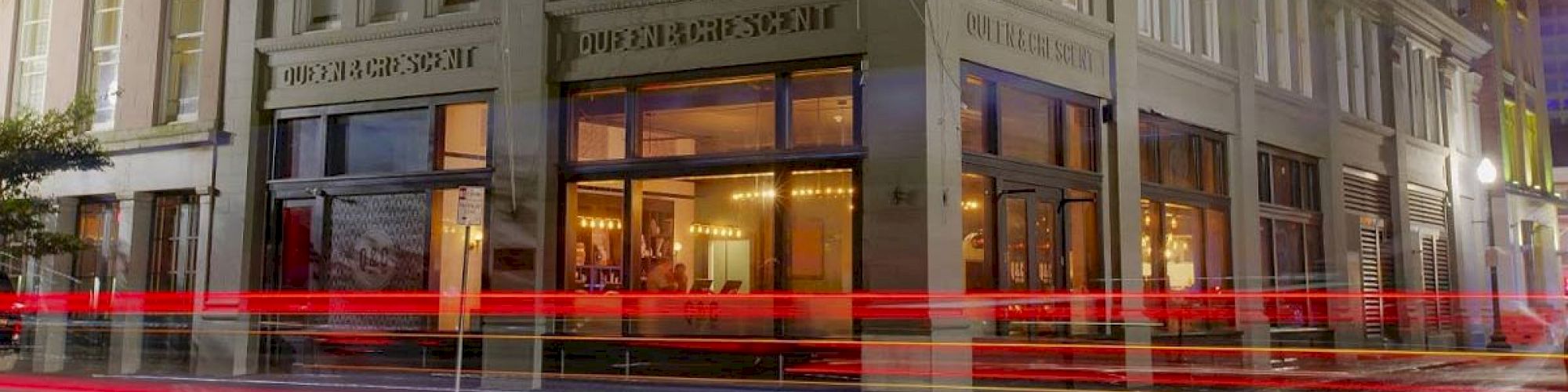 An image of the exterior of a building with "QUEEN & CRESCENT" on its facade, showing light trails from passing vehicles in front of it.