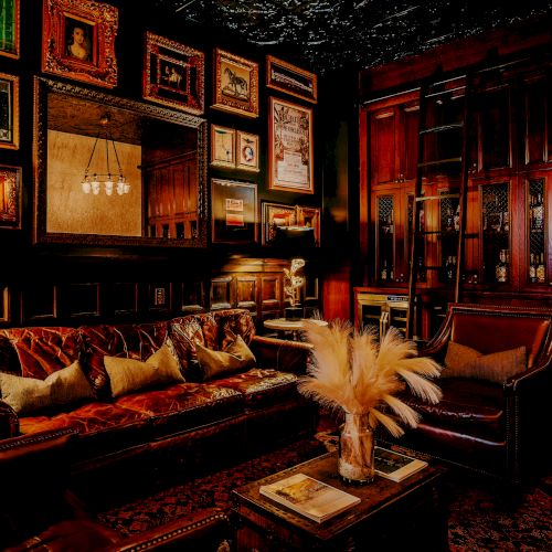 A cozy, darkly lit living room with plush leather seating, framed artwork on the walls, a chandelier, and a glass-front cabinet showcasing decor. Sentence ends here.