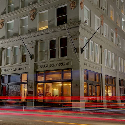 A nighttime image of a corner building with illuminated windows and streaks of red lights from passing cars.