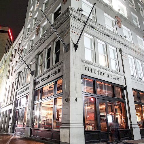 A nighttime street view. The scene features a brightly illuminated corner building with large windows, housing 