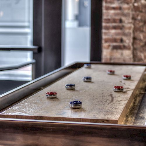 The image shows a shuffleboard table with various colored pucks arranged on it, set in a room with brick walls and large windows.