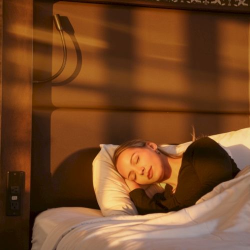 A person is sleeping on a bed in a dimly lit hotel room, head resting on a pillow, across a white duvet, with decorative wallpaper and a headboard behind. Always ending the sentence.