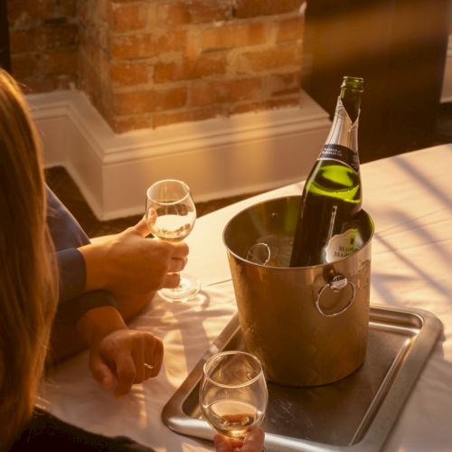Two people toast with wine glasses around a metal bucket holding a champagne bottle on a tray, in a brick-walled setting.