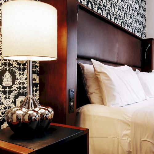 A stylish hotel bedroom with a patterned wallpaper, a tall lamp on a dark wood nightstand, and a white, neatly made bed against a wooden headboard.