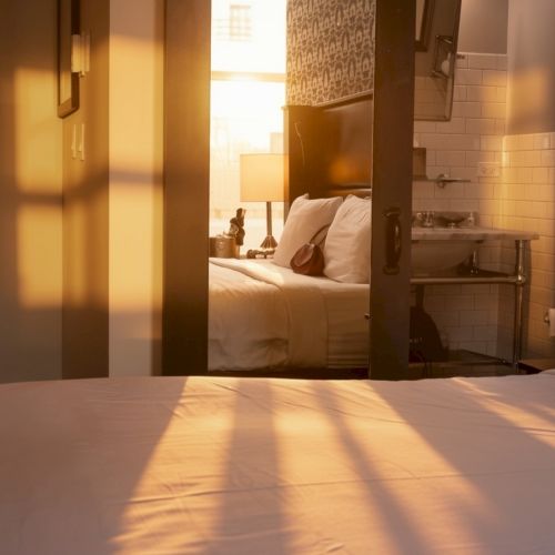 A cozy hotel room seen through a doorway: warm sunset light on a white bed, a small bedside area with a lamp, and a softly lit bathroom in the background.