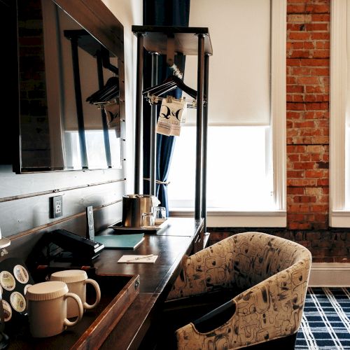A hotel room workspace with a desk, coffee mugs, a hanging garment rack, a patterned chair, and a brick wall beside a window, in warm lighting.
