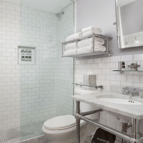 A modern bathroom with a glass-enclosed shower, white subway tiles, a toilet, a sink with a mirror, and towel storage above the toilet.