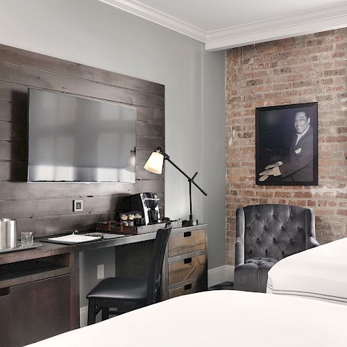 The image depicts a hotel room with a modern rustic design, featuring a wall-mounted TV, a desk with a lamp, a chair, and two beds.