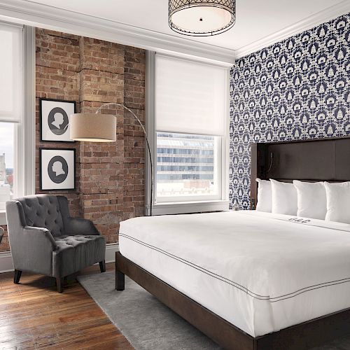 A modern bedroom with a king-size bed, patterned wallpaper, brick accent wall, grey armchair, and wall art, featuring large windows and wooden flooring.