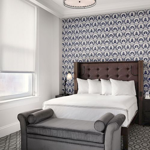 A stylish bedroom features a bed with a tufted headboard, white bedding, and four pillows, a patterned accent wall, a gray bench at the foot, and bedside tables.