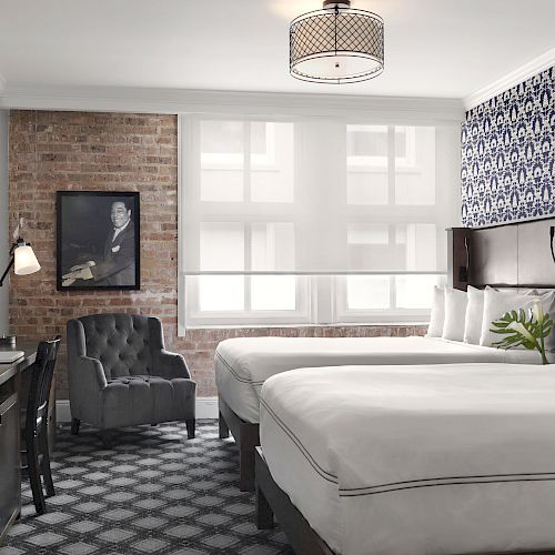 A modern hotel room with two white beds, a patterned accent wall, brick wall, grey armchair, TV on a wooden desk, and a window bringing in light.