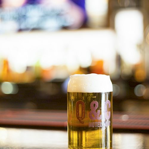 A glass of beer with a frothy head sits on a bar counter, with a blurred background of lights and indistinct figures.