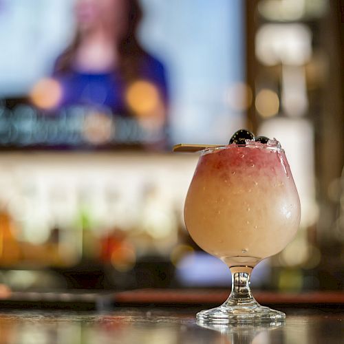 The image shows a fancy cocktail with layers of red and white served in a round glass. The background is a blurred bar and a TV screen.