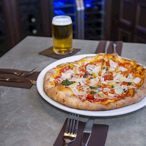 A pizza on a plate, a glass of beer, a lit candle, and utensils are set on a restaurant table.