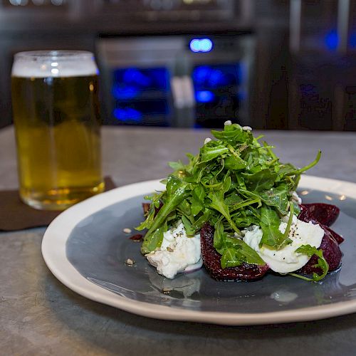 A plate with a salad of greens and other ingredients alongside a glass of beer on a table with a candle in the background.