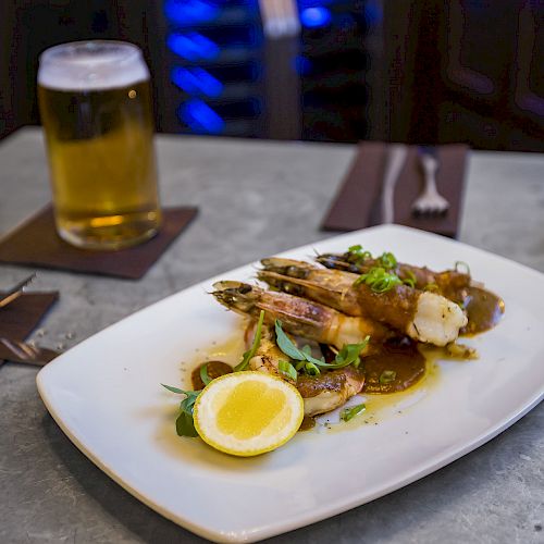 A plate of shrimp with garnish and a lemon slice, accompanied by a beer and candle in a cozy dining setting.