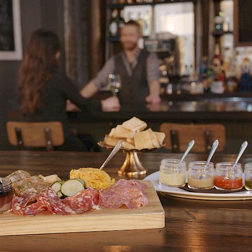 A charcuterie board with meats, cheeses, and jars of spreads is on a bar counter. There are two people chatting in the background.