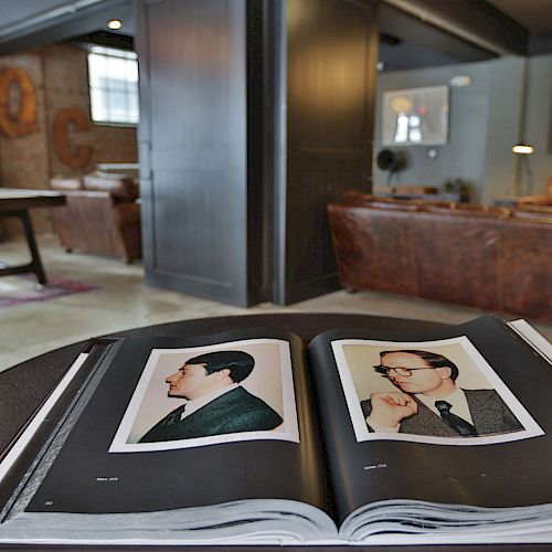 An open book with portraits is placed on a table in a modern, cozy lounge area with leather couches and industrial decor.