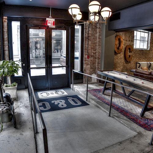 A modern interior features a glass door entrance, potted plants, a rug, and a table. The space has brick walls and industrial decor, including wall art.