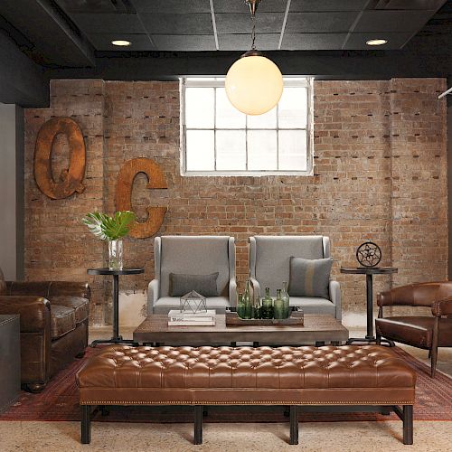 A cozy living room with brick walls, leather seating, two armchairs, a tufted bench, a round light fixture, and decorative wall letters.