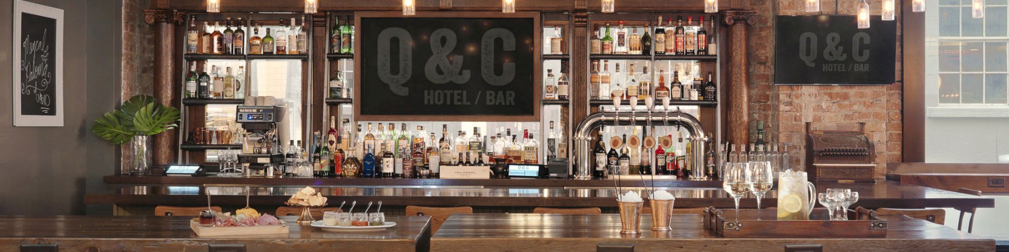 A stylish bar with wooden high chairs, pendant lights, and a well-stocked bar area. The sign "O&C Hotel & Bar" is visible in the background.