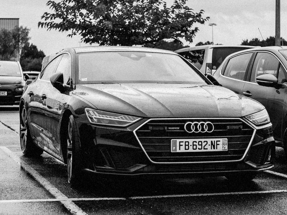 A black-and-white photo of an Audi parked in a lot, featuring a clear view of the front grille and license plate.