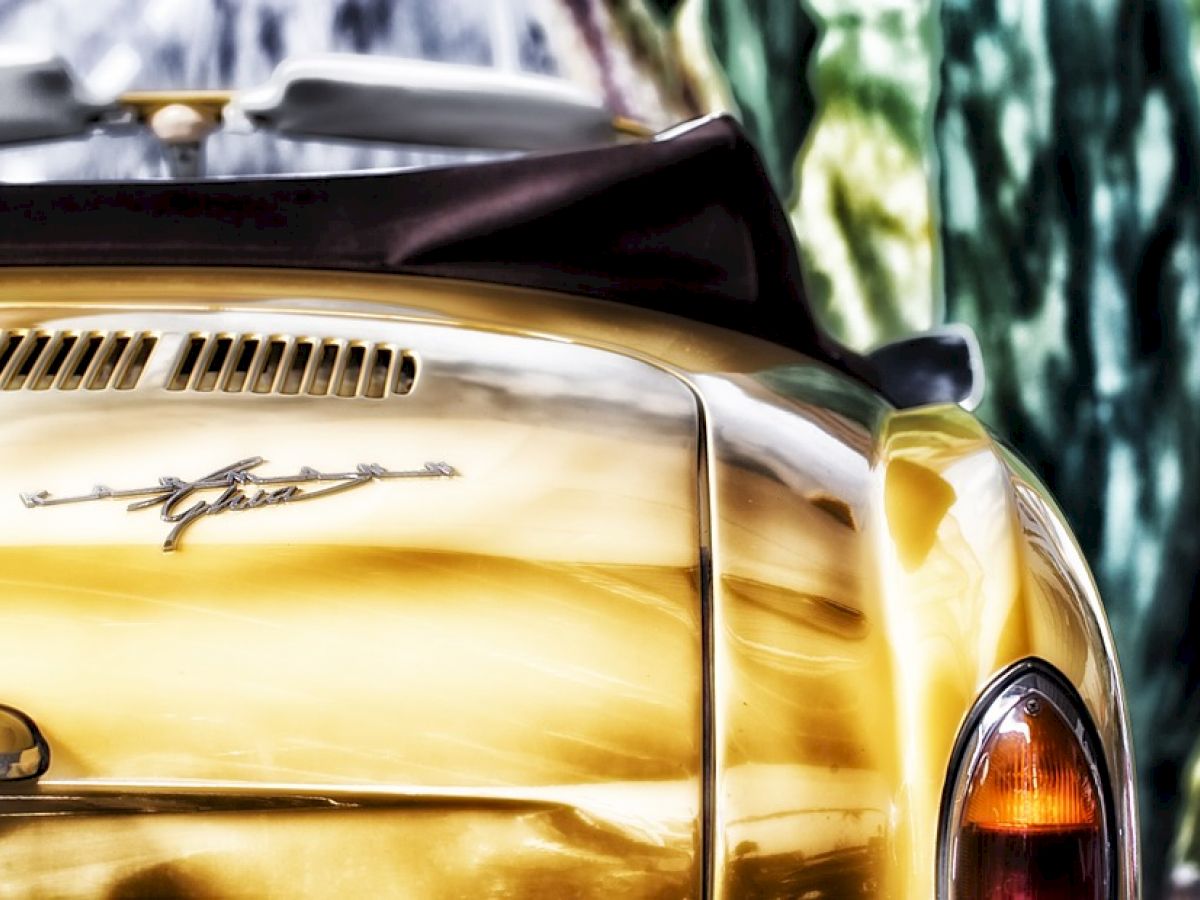 The image shows the rear of a yellow vintage convertible car with a distinctive emblem and a blurred natural background.