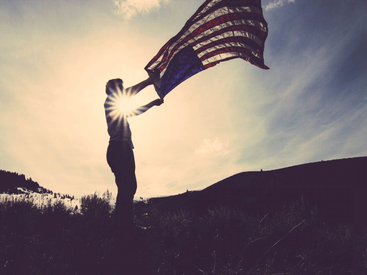 A silhouette of a person holding an American flag against a bright sky, with the sun creating a starburst effect.