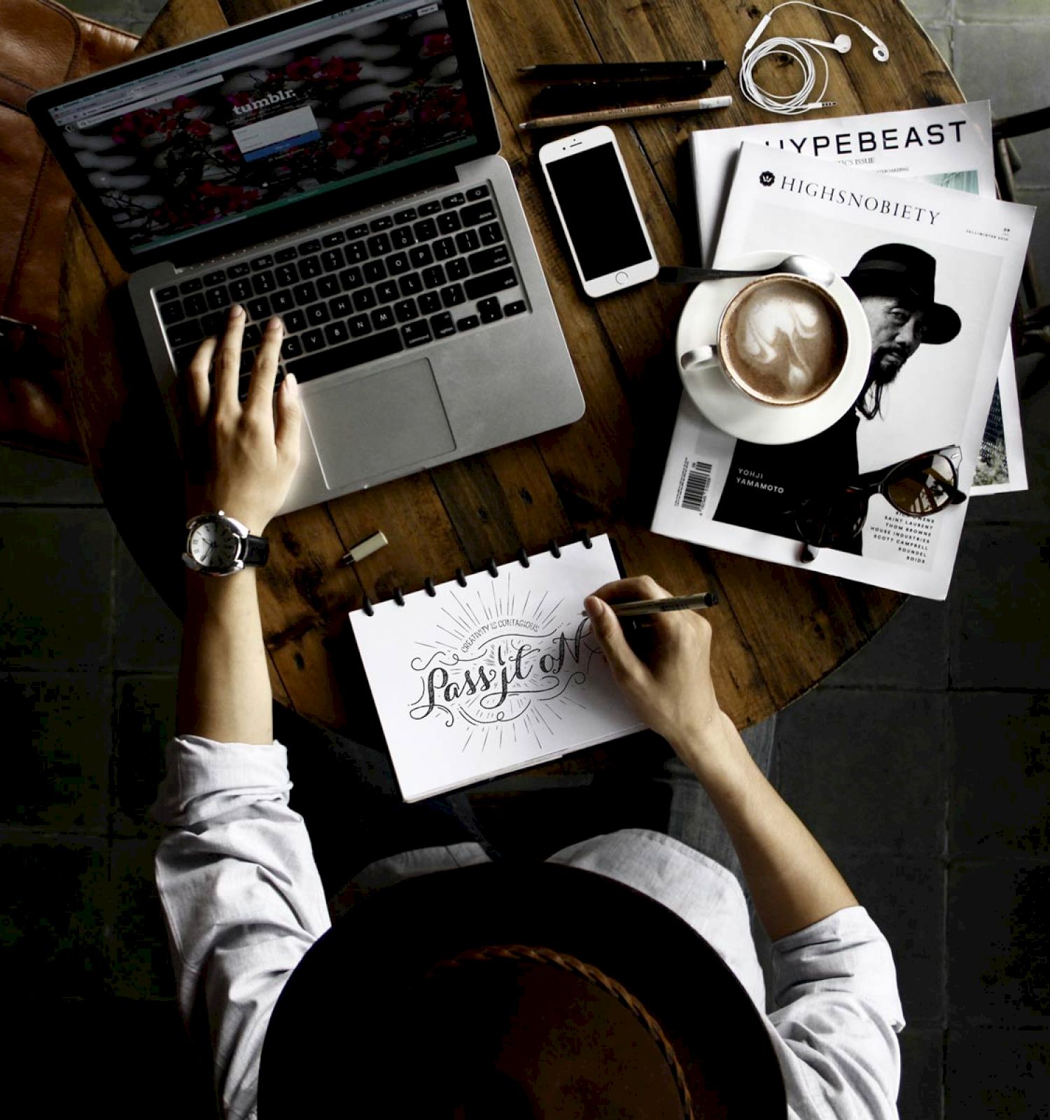 A person is working at a wooden table with a laptop, phone, coffee, magazines, and a handwritten note that says "Focused." The view is from above.