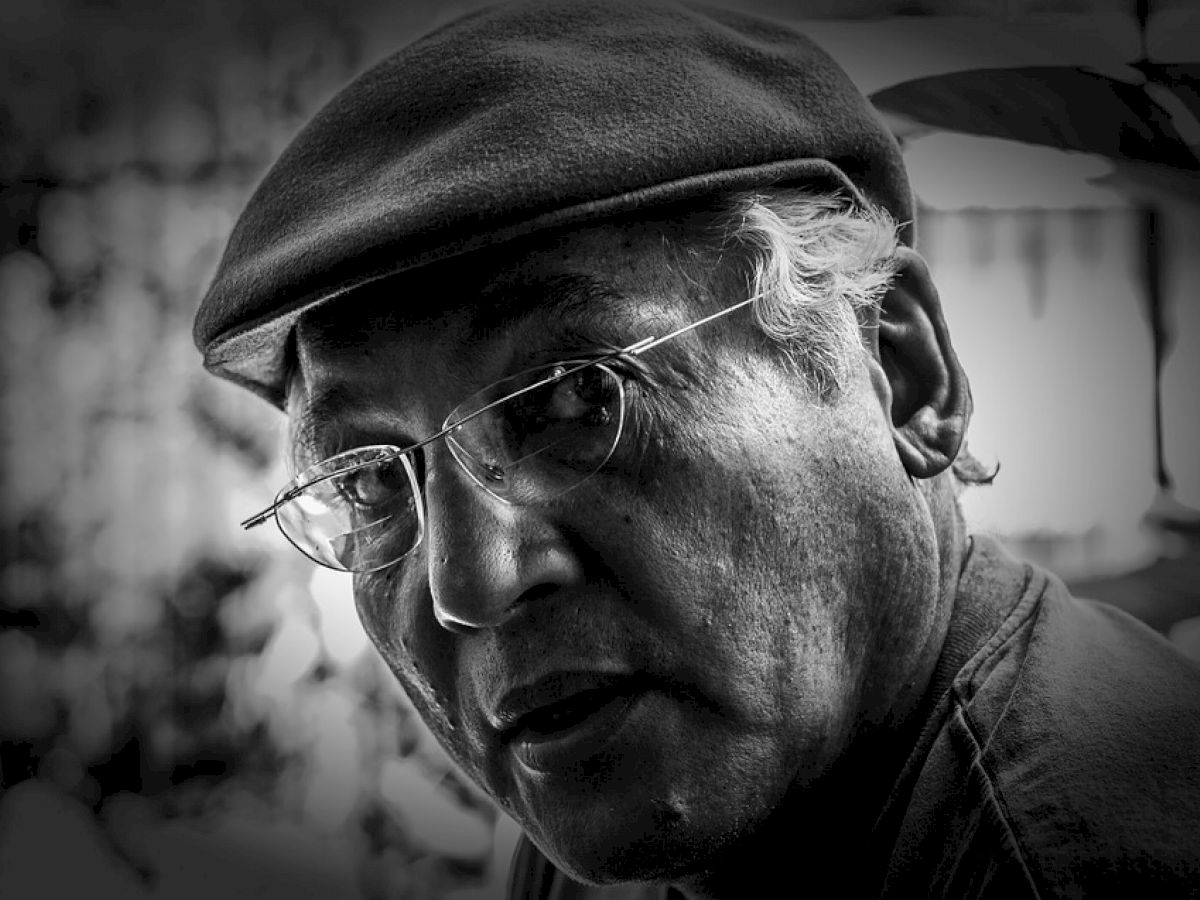 A black and white photo of an older man wearing a cap and glasses, with a thoughtful expression and a blurred background ending the sentence.