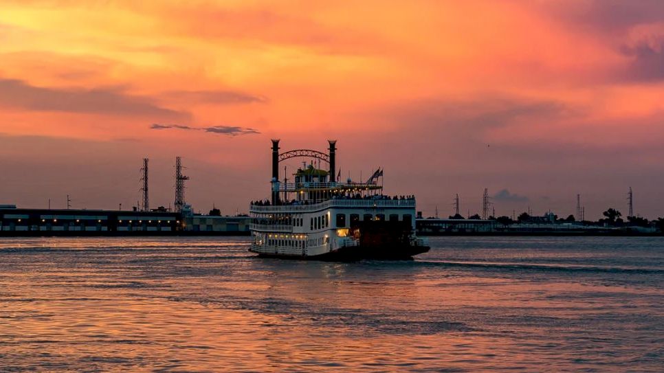 A riverboat cruises on a calm waterbody with a beautiful sunset casting orange and pink hues across the sky in the background.