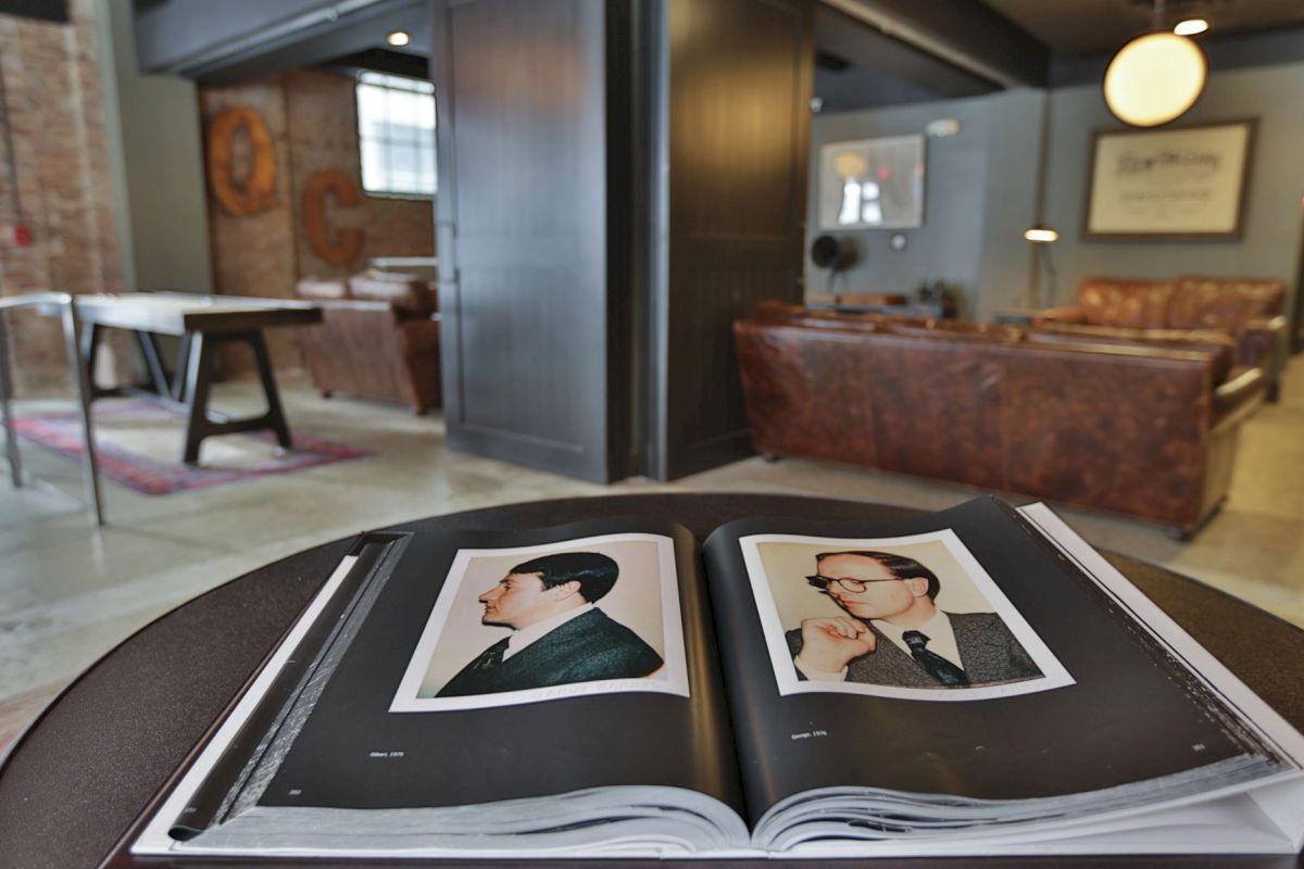 The image shows an open book on a table with photos of two men on its pages in a stylishly furnished room with leather couches and a projector screen.