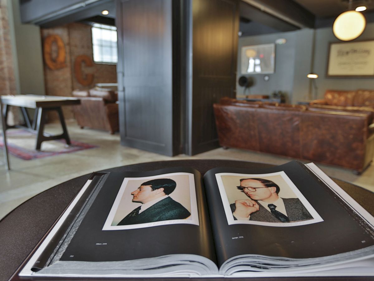 The image shows an open book on a table with photos of two men on its pages in a stylishly furnished room with leather couches and a projector screen.