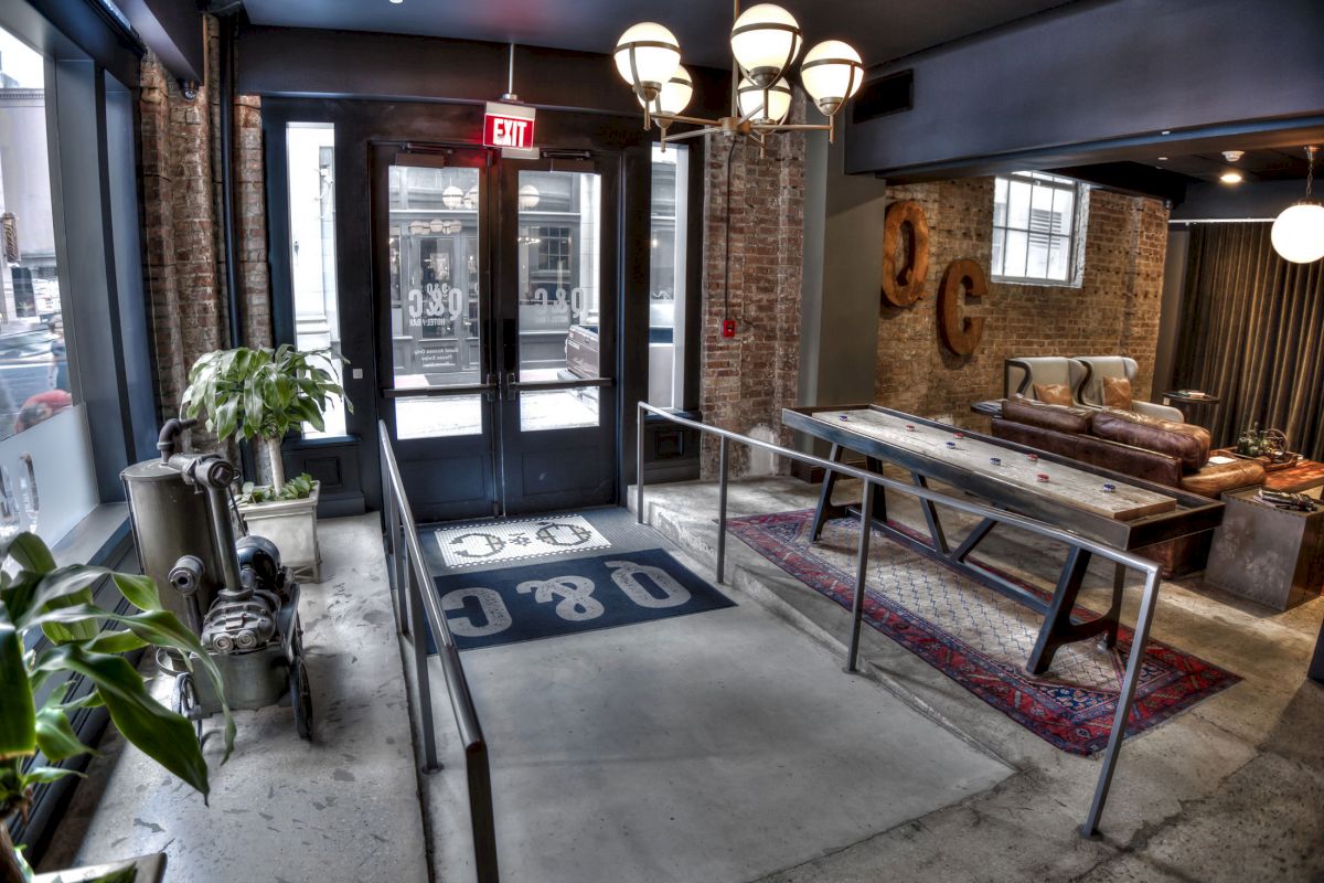 The image shows a stylish interior with a ramp leading to a double door, plants, industrial decor, and a seating area with a rug and couch.