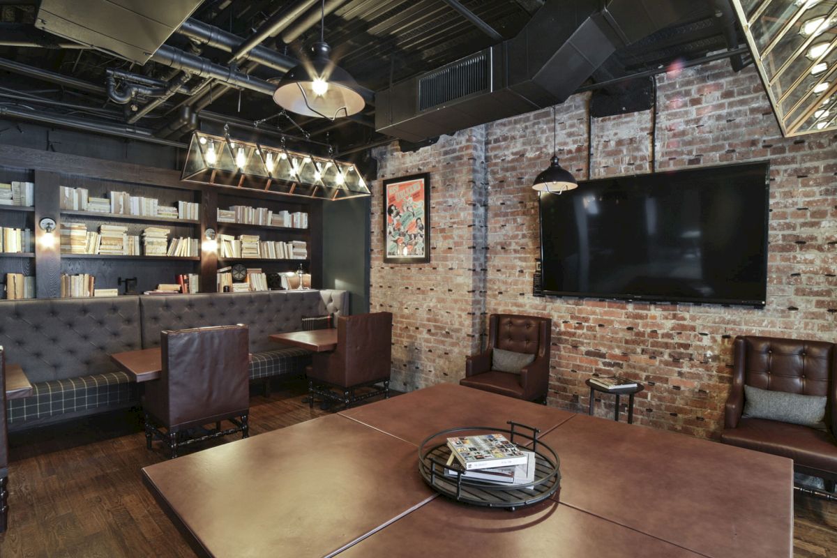 A cozy room with a brick wall, shelves of books, leather chairs, a large TV, pendant lights, and a wooden table with books and magazines.