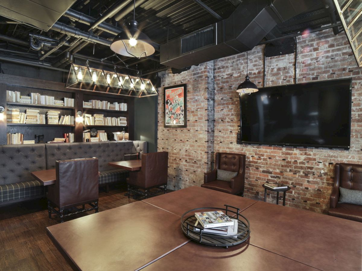 A cozy room with a brick wall, shelves of books, leather chairs, a large TV, pendant lights, and a wooden table with books and magazines.