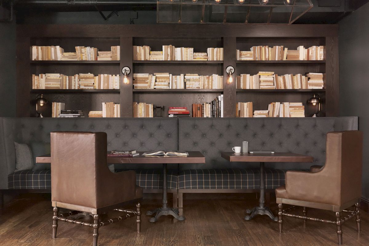 A cozy library-themed cafe with a dark, tufted sofa, wooden tables, leather chairs, and bookshelves filled with books on the wall.